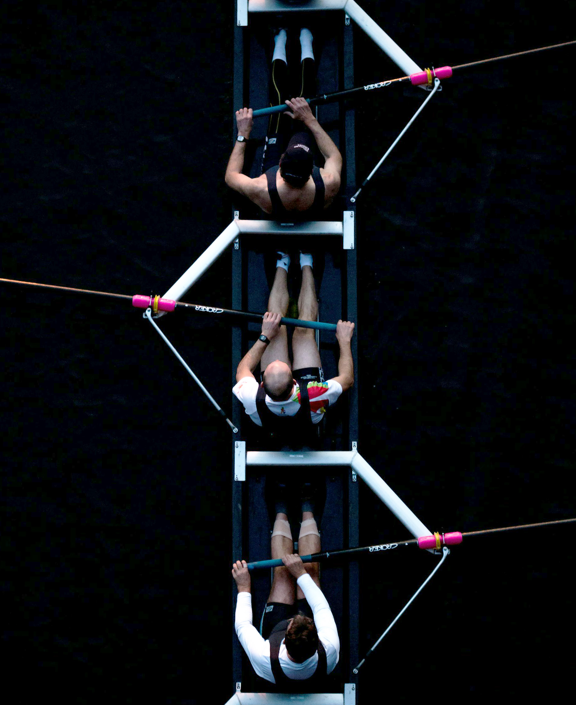 Aerial view of three male rowers in a boat, rowing in sync on dark water.