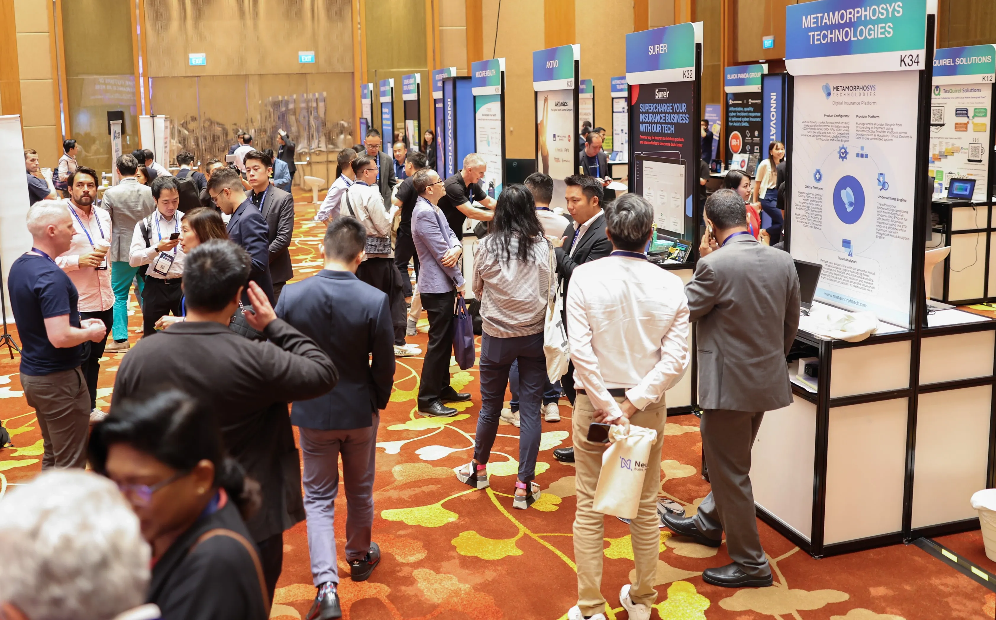 Attendees networking and visiting exhibition booths at a professional conference in a large, carpeted hall.