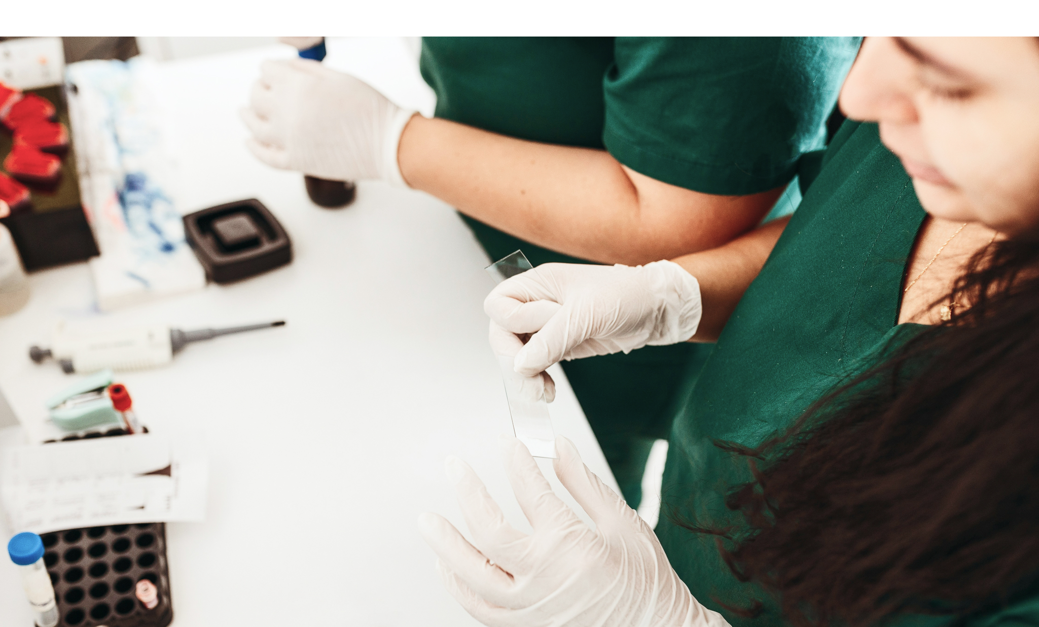Two healthcare professionals in green scrubs and gloves examining a microscope slide on a white lab table.