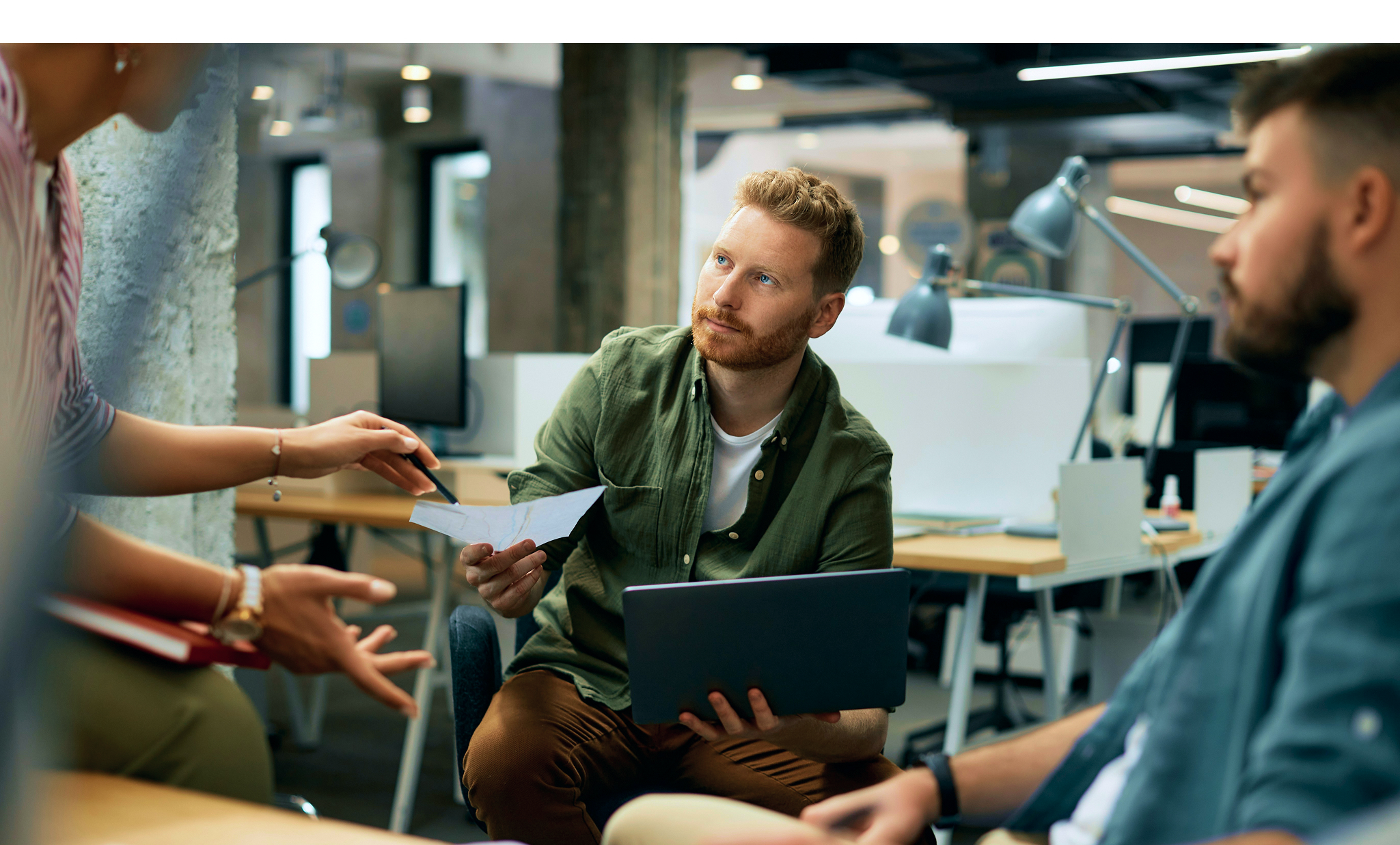 Three colleagues in a modern office engaged in a discussion, one holding a laptop and another pointing at a paper.