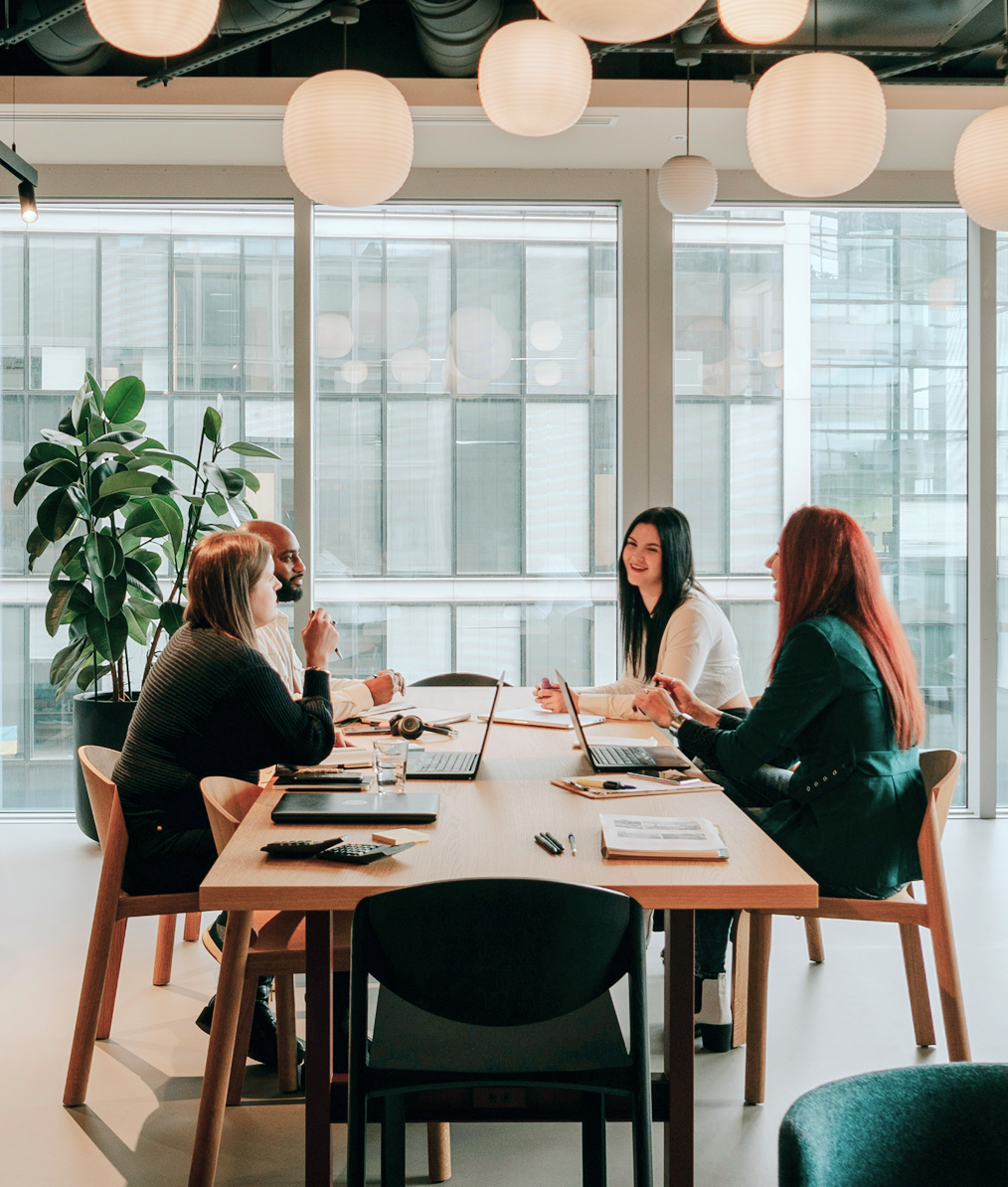Four diverse professionals engaged in a meeting around a table in a modern office with large windows and hanging lamps.